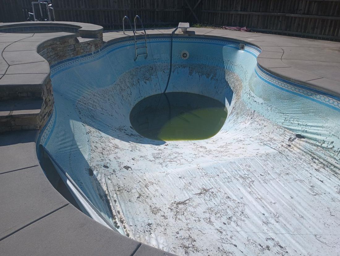 Kidney-shaped pool drained, with a faded, debris-covered old liner and green water in the deep end